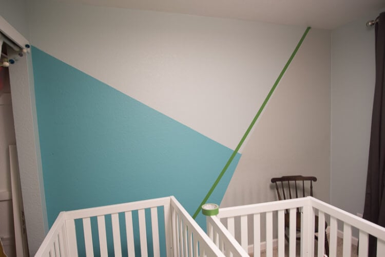Two baby cribs and a rocking chair next to a wall marked off with green painter\'s tape and lower part painted in aqua