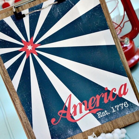 Vintage patriotic 4th of July themed sign clipped to a clip board that is sitting on a table propped up next to a red lantern and red and white pom poms