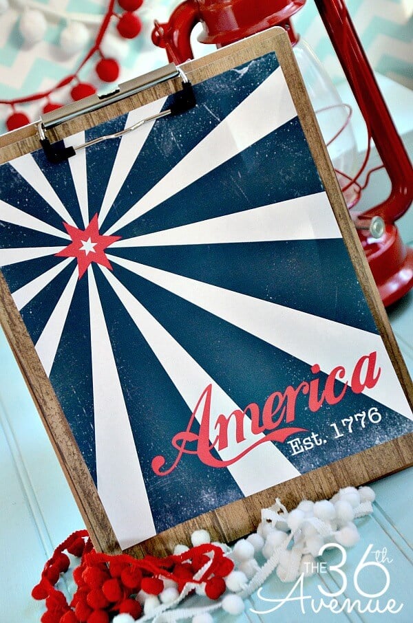 Vintage patriotic 4th of July themed sign clipped to a clip board that is sitting on a table propped up next to a red lantern and red and white pom poms