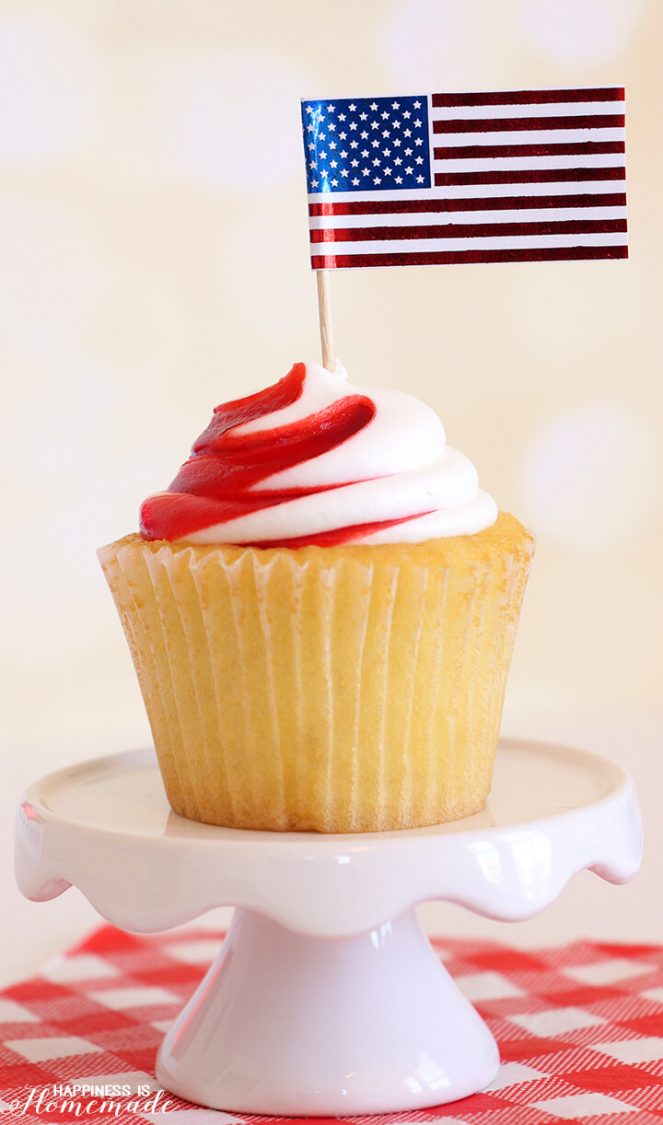 A cupcake on top of a white pedestal, that has white and red frosting on it and a foiled flag cupcake topper