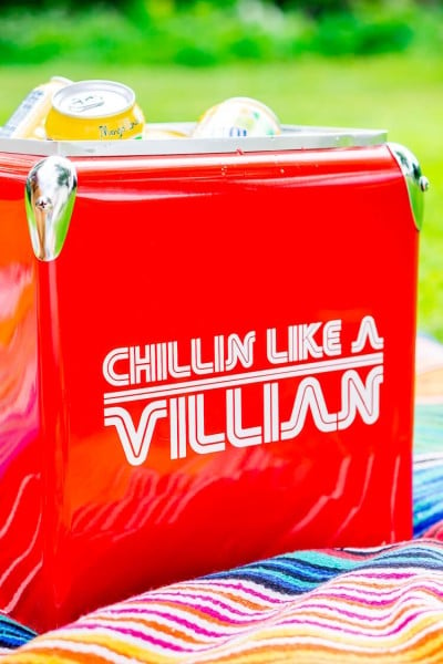A red ice cooler sitting on top of a striped blanket and filled with beverages and a sign on the side of the cooler that says, "Chillin Like a Villian"