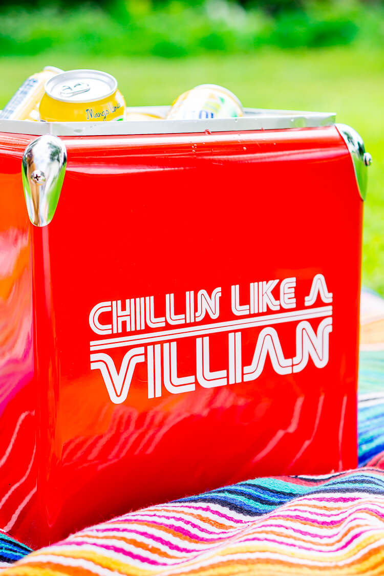 A red ice cooler sitting on top of a striped blanket and filled with beverages and a sign on the side of the cooler that says, \"Chillin Like a Villian\"