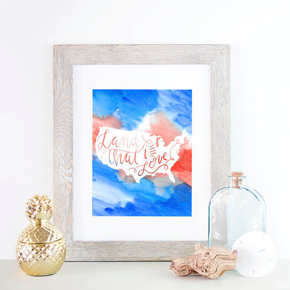 A gray colored frame of a patriotic picture of an outline of the USA that says, \"Land That We Love\" sitting on a table next to gold and glass decor

