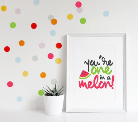 A plant sitting on a table next to a colorful polka dotted wall and a white framed picture against the wall that has a piece of watermelon on it and text that says, \"You\'re One in a Melon!\"