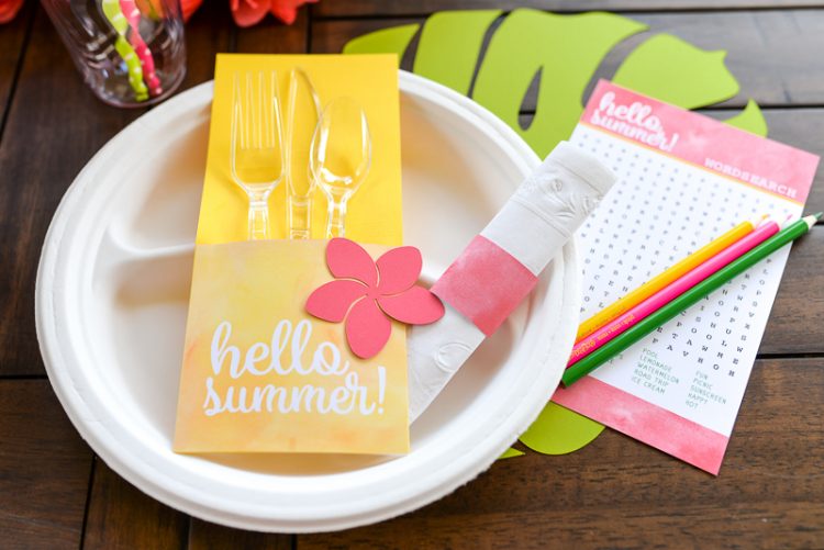 A table with fruit, flowers and a place setting that contains plastic cutlery wrapped in a utensil holder made from paper cardstock and pencils on top of a word serach puzzle