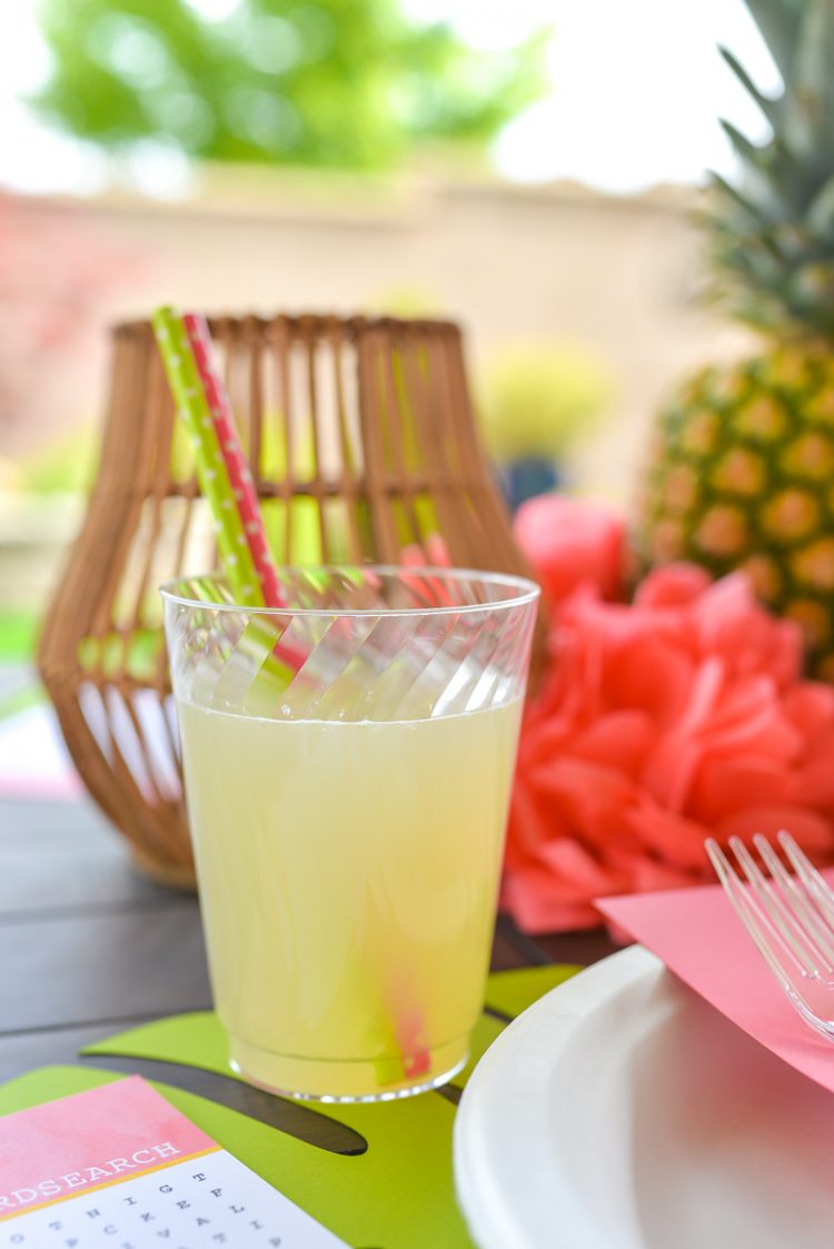 A close up of a glass with two straws in it next to a place setting, a pineapple, a candle and paper vcut flowers