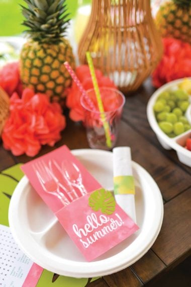 A table with fruit, flowers and a place setting that contains plastic cutlery wrapped in a utensil holder made from paper cardstock