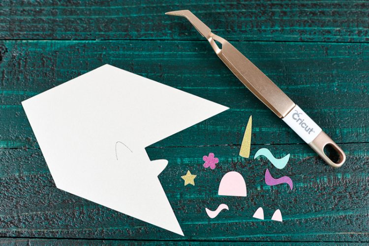 A Cricut tweezers and pieces of a unicorn bookmark sitting on top of a dark green table