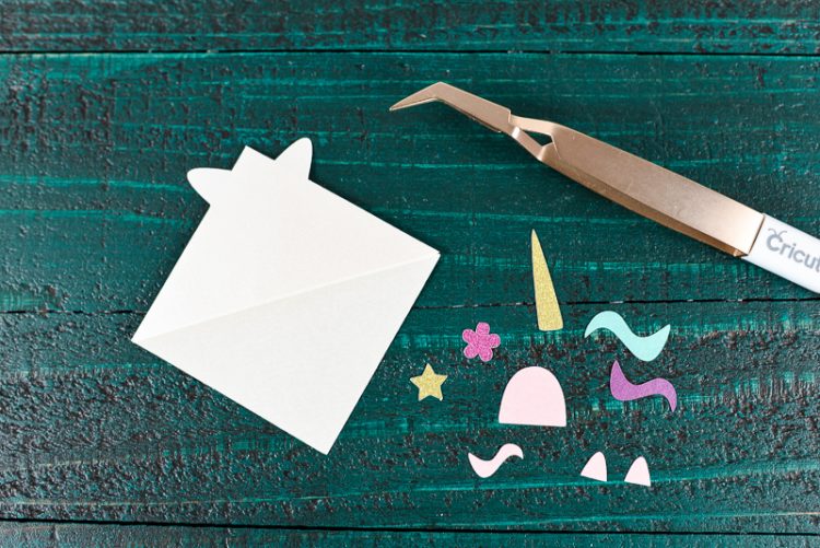 A Cricut tweezers and pieces of a unicorn bookmark sitting on top of a dark green table