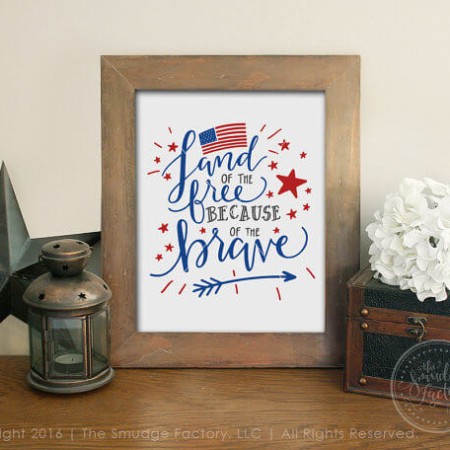 A wooden framed picture sitting on a table next to some tin style decor and the picture has a USA flag on it and the text. "Land of the Free Because of the Brave"