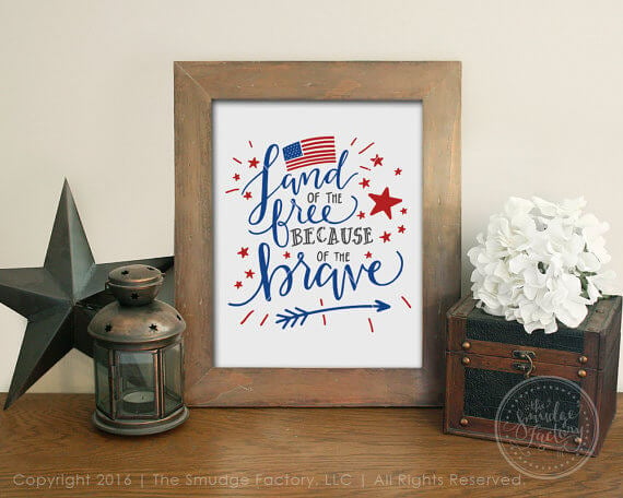 A wooden framed picture sitting on a table next to some tin style decor and the picture has a USA flag on it and the text. \"Land of the Free Because of the Brave\"