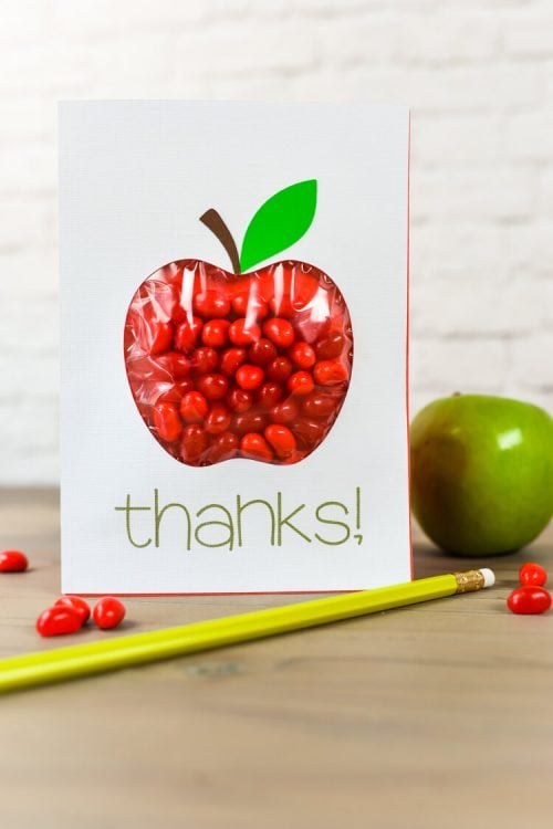 A green apple, a pencil and candy sitting on a table along with a card that says, "Thanks" and is decorated with an apple image and filled with red candy