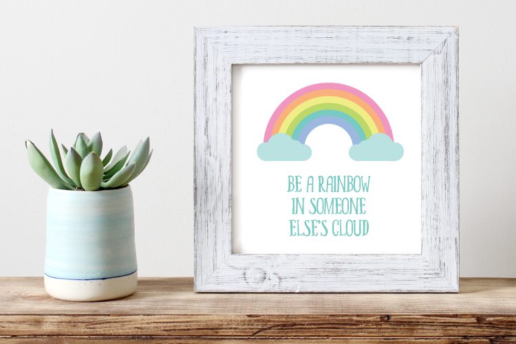 A potted succulent sitting on a table along with a weathered white framed picture that has a pastel-colored rainbow with clouds on it and the saying, \"Be a Rainbow in Someone Else\'s Cloud\"