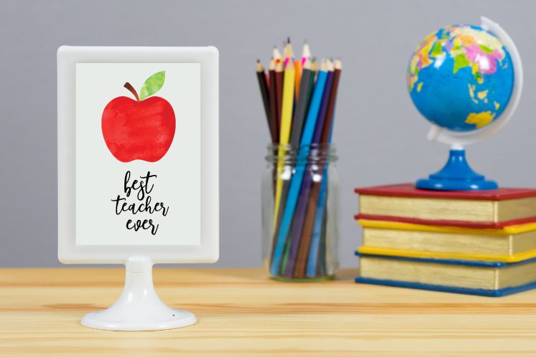 On a desk is sitting a jar of colored pencils, a small world globe sitting on top of three books and a pedestal white frame with a picture of an apple on it and the saying, \"Best Teacher Ever\"