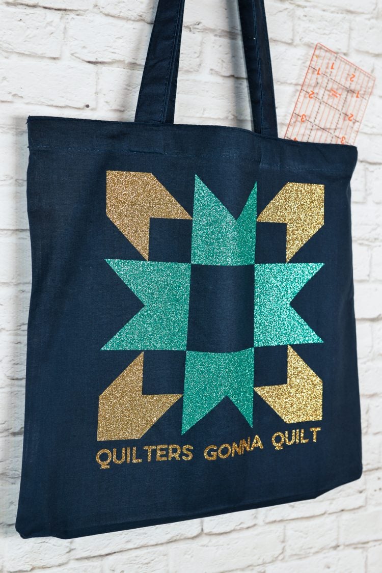 Close up of a black canvas tote hanging from a white brick wall and the tote is decorated with an aqua and a gold-colored quilt block image and the saying, \"Quilters Gonna Quilt\"
