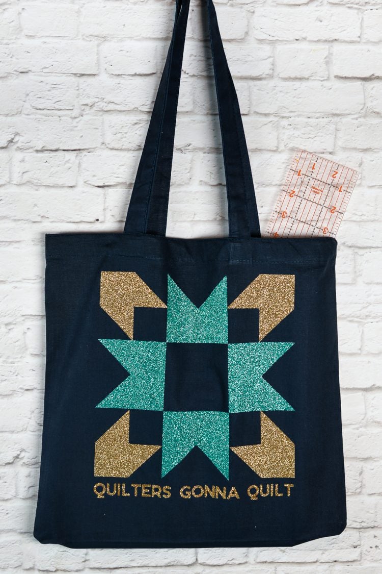 Close up of a black canvas tote hanging from a white brick wall and the tote is decorated with an aqua and a gold-colored quilt block image and the saying, \"Quilters Gonna Quilt\"