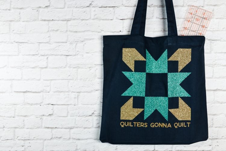 A black canvas tote hanging from a white brick wall and the tote is decorated with an aqua and a gold-colored quilt block image and the saying, \"Quilters Gonna Quilt\"