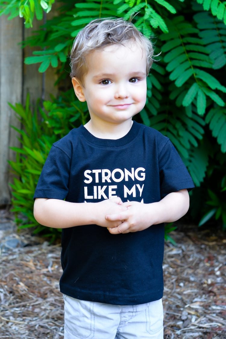 A little boy wearing a dark blue t-shirt that says, \"Strong Like My Mom\"