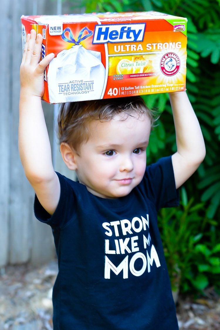 A little boy holding box of Hefty Ultra Strong garbage bags on his head and wearing a dark blue t-shirt that says, \"Strong Like My Mom\"