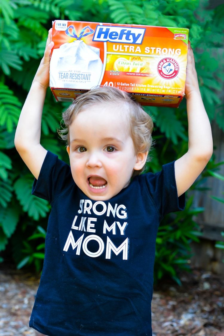 A little boy holding box of Hefty Ultra Strong garbage bags on his head and wearing a dark blue t-shirt that says, \"Strong Like My Mom\"
