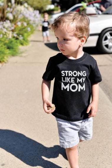 A little boy walking outside, wearing blue jean shorts and a black t-shirt that says, "Strong Like My Mom"