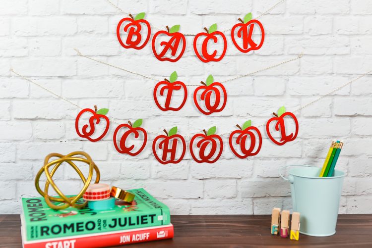 Books, a bucket of pencils and other items sitting on a desk that has a banner with cut out apples hanging above it that says, \"Back to School\"