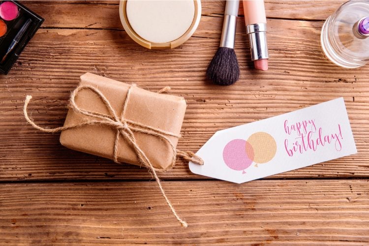 Makeup sitting on a table along with a package wrapped in brown paper and tied with twine and has a gift tag attached that says, \"Happy Birthday\"