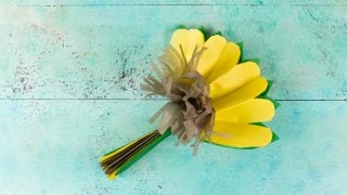 On an aqua blue table lays brown, yellow and green tissue paper folded together and partially pulled apart to resemble a sunflower