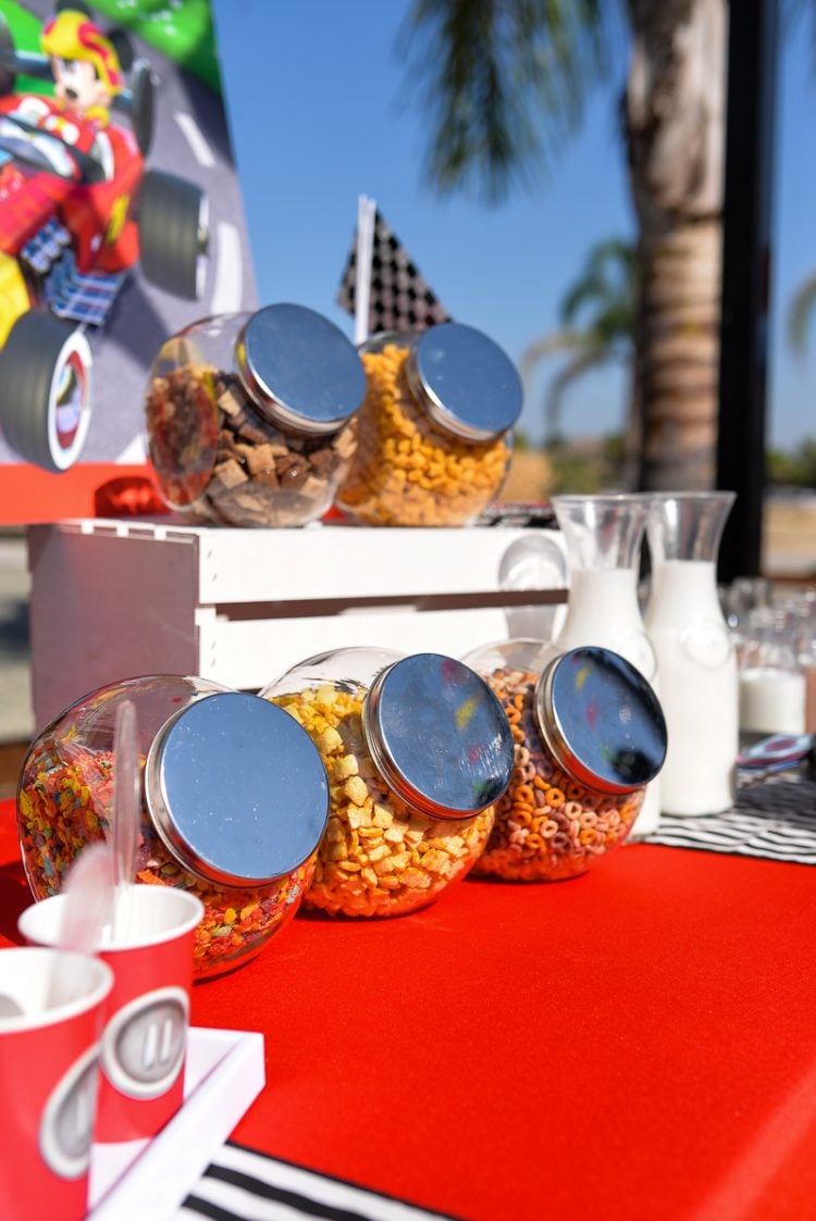 A table with a red tablecloth with jars filled with cereal and two carafes filled with milk