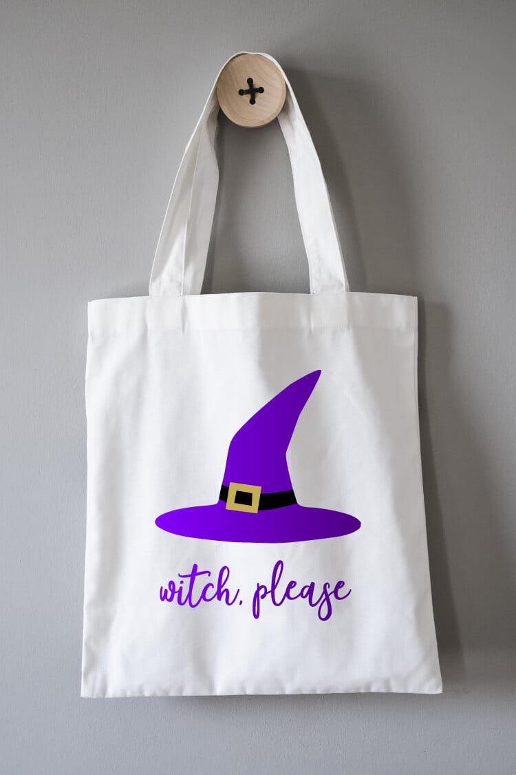 A white canvas tote hanging from a hook, and the tote is decorated with a witch\'s hat and the text, \"Witch, Please\"