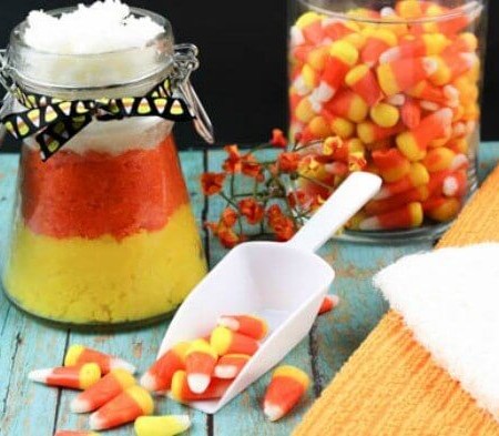 Sitting on an aqua blue table is a jar filled with cand corn candy, a white scoop with candy corn candy spilling out of it and another jar decorated a yellow, orange and white substance