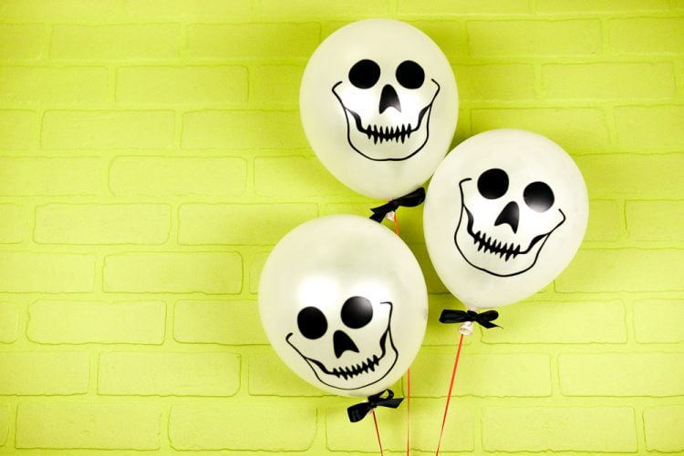 Against a lime green wall are three white balloons decorated with skulls