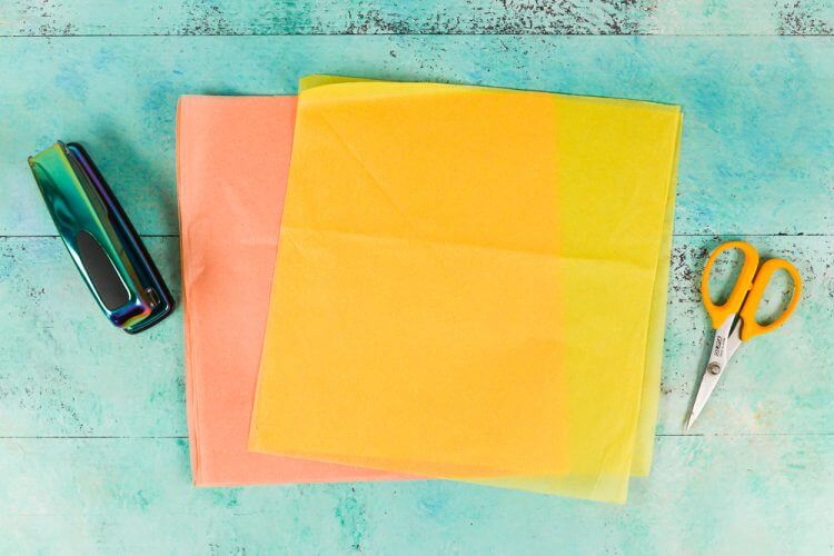 A stapler, scissors and a piece of yellow and a piece of orange tissue paper lying on an aqua blue table