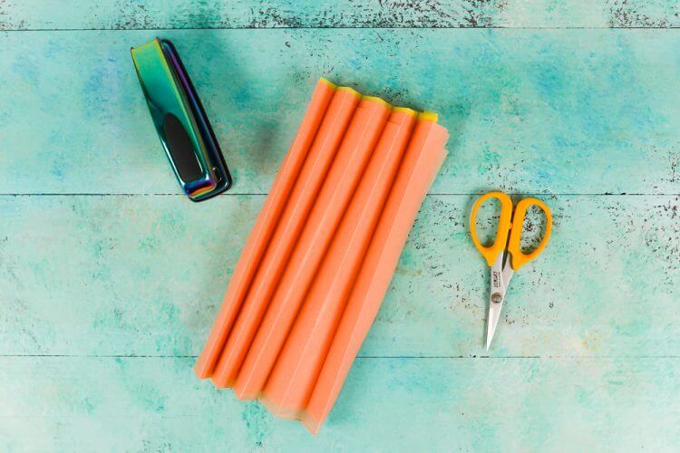 A stapler, scissors and a piece of orange accordion folded tissue paper lying on an aqua blue table