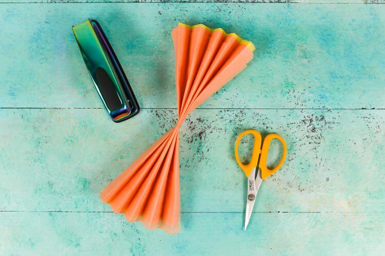A stapler, scissors and a piece of orange accordion folded tissue paper lying on an aqua blue table