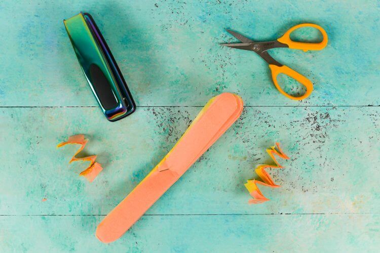 A stapler, scissors and a piece of orange folded tissue paper lying on an aqua blue table