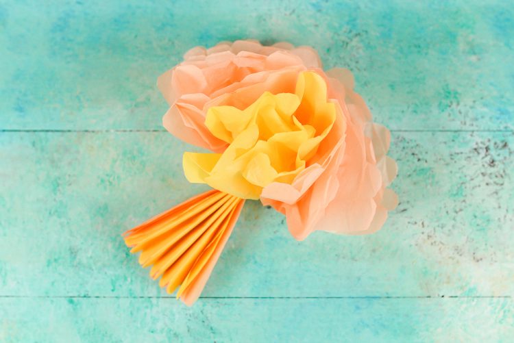 Lying on an aqua blue table is an orange and yellow accordion folded tissue paper where part of the tissue paper is pulled apart to resemble a flower