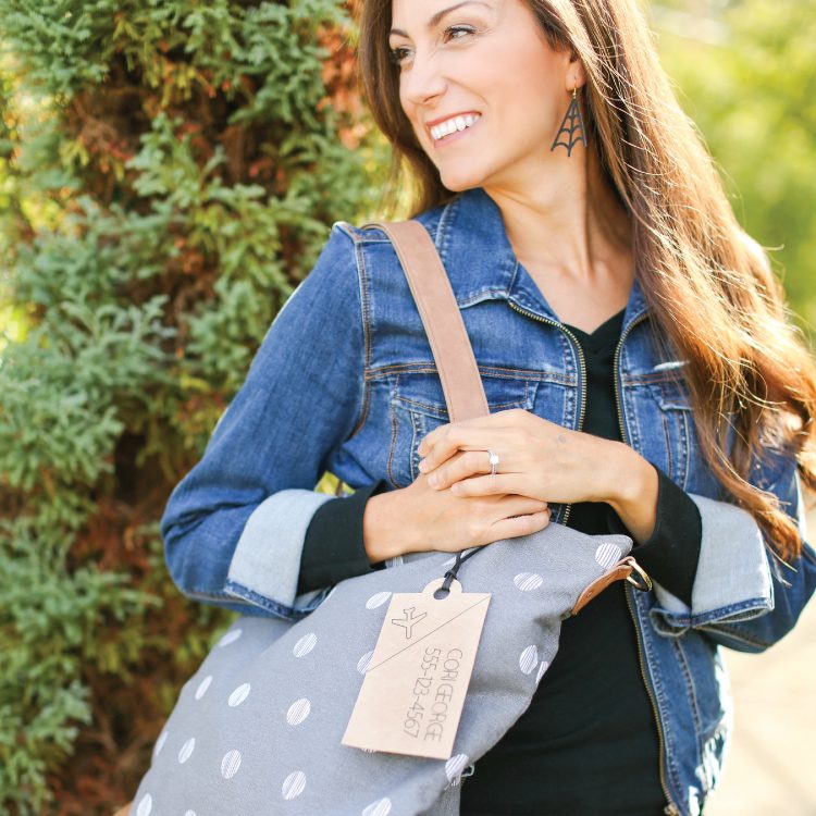 A woman standing outside in a blue jean jacket, a black shirt, an earring and holding a large gray tote bag