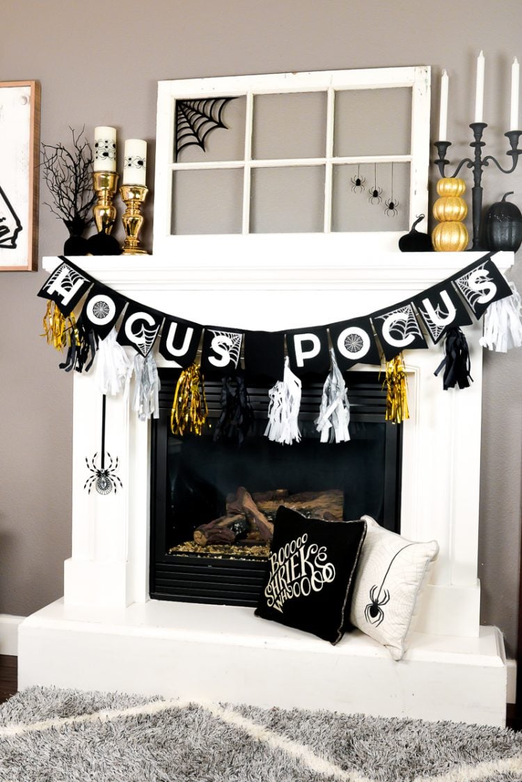 Halloween black, white and gold decor adorning a white fireplace that includes, gold and black pumpkins, white candles in black candelabras and a banner that says, \"Hocus Pocus\"