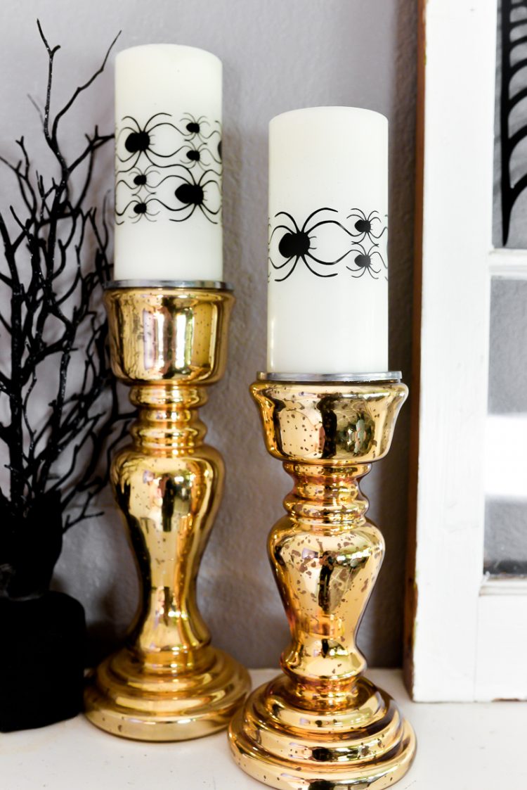 A close up of two gold candlesticks with white candles decorated with images of spiders