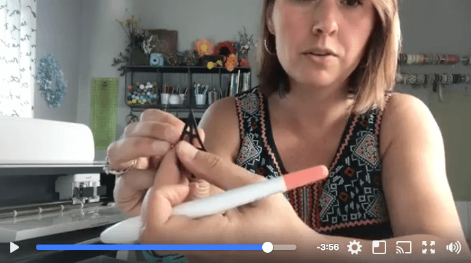 A Cricut machine on a table where a woman is sitting and holding a Cricut weeding tool and a piece of a cut out material