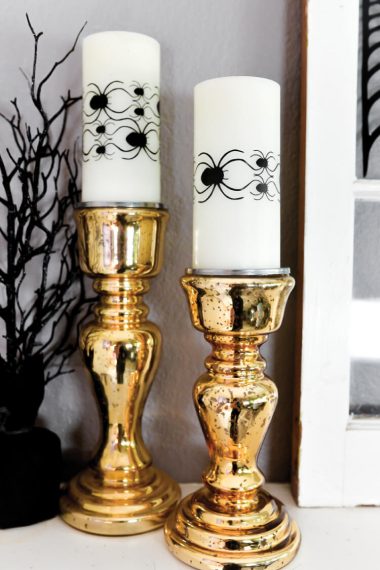 Sitting on a table are two gold candlesticks with white candles decorated with images of spiders