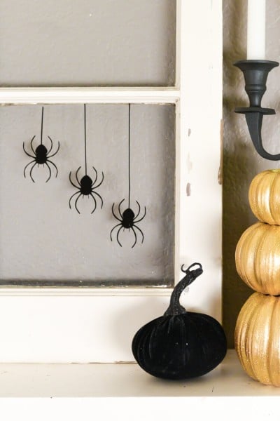 Close up of an old window decorated with hanging black spiders sitting on top of a fireplace mantel and next to candles and pumpkins