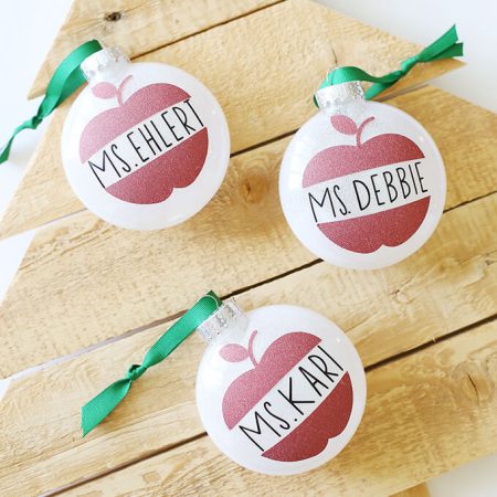 Three white ornaments laying on top of a wooden tree, and ornaments are decorated with an apple and personalized with a name