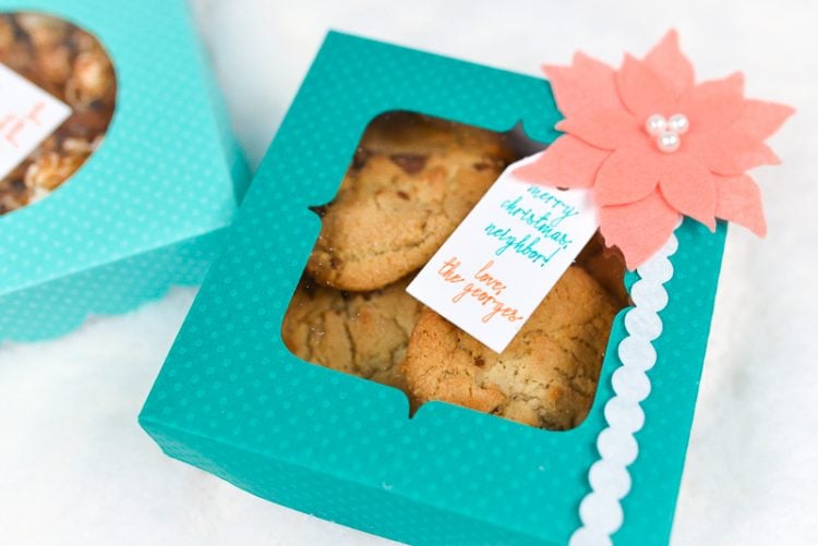 An aqua colored polka dot box filled with cookies and decorated with a coral colored felt poinsettia and a gift tag 