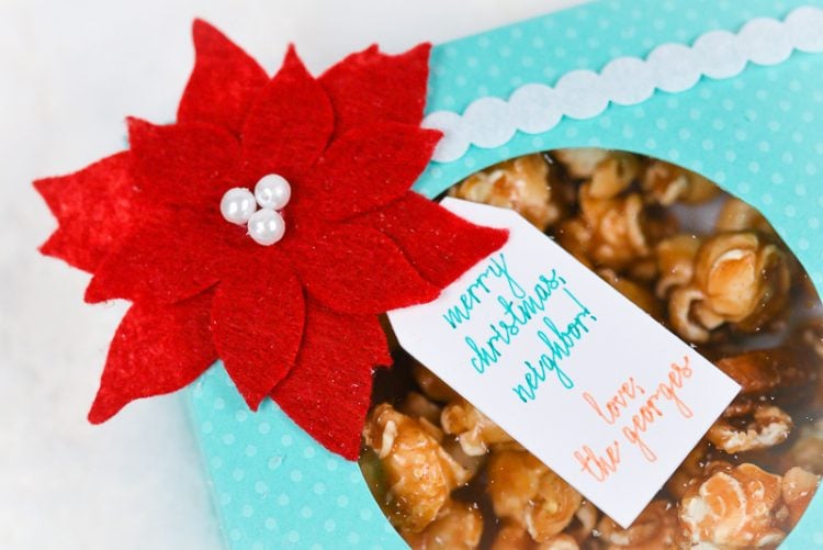 An aqua colored polka dot box filled with caramel popcorn and decorated with a red felt poinsettia and a gift tag 