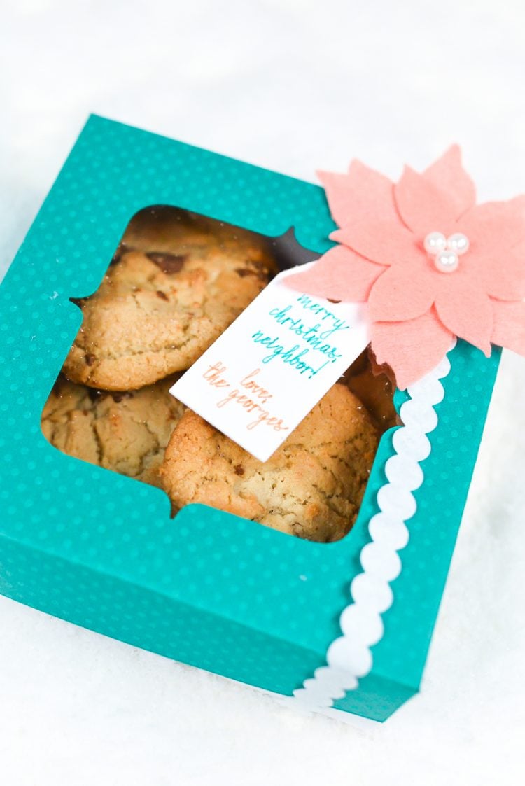 An aqua colored polka dot box filled with cookies and decorated with a coral colored felt poinsettia and a gift tag 