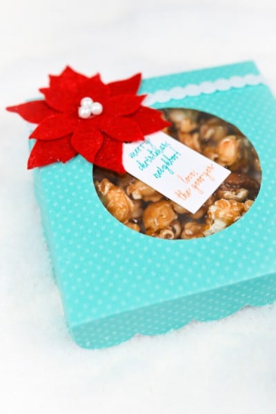 An aqua colored polka dot box filled with caramel popcorn and decorated with a red felt poinsettia and a gift tag
