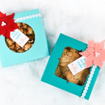 Two aqua colored polka dot boxes filled with cookies and caramel popcorn with one decorated with a coral colored felt poinsettia and a gift tag and the other decorated with a red felt poinsettia and a gift tag