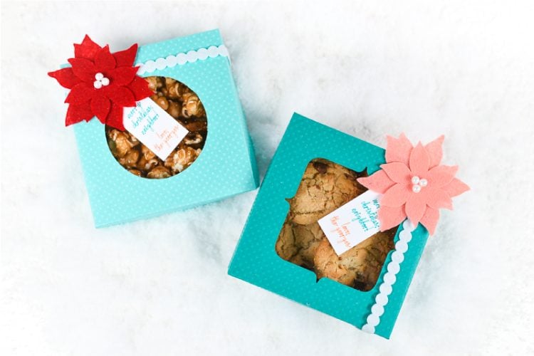 Two aqua colored polka dot boxes filled with cookies and caramel popcorn with one decorated with a coral colored felt poinsettia and a gift tag and the other decorated with a red felt poinsettia and a gift tag